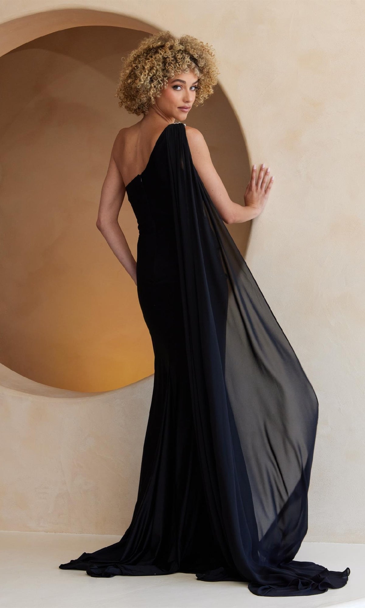 A woman with curly hair models the Amelia Couture 7056, a one-shoulder long prom dress with a sheer drape, standing before a beige wall with a circular cutout.